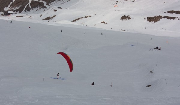 Anthony Green skis over a line in the snow, before getting airborne again and completing the course for good points. The glider on the right is flying along a line of sticks, kicking each one. 